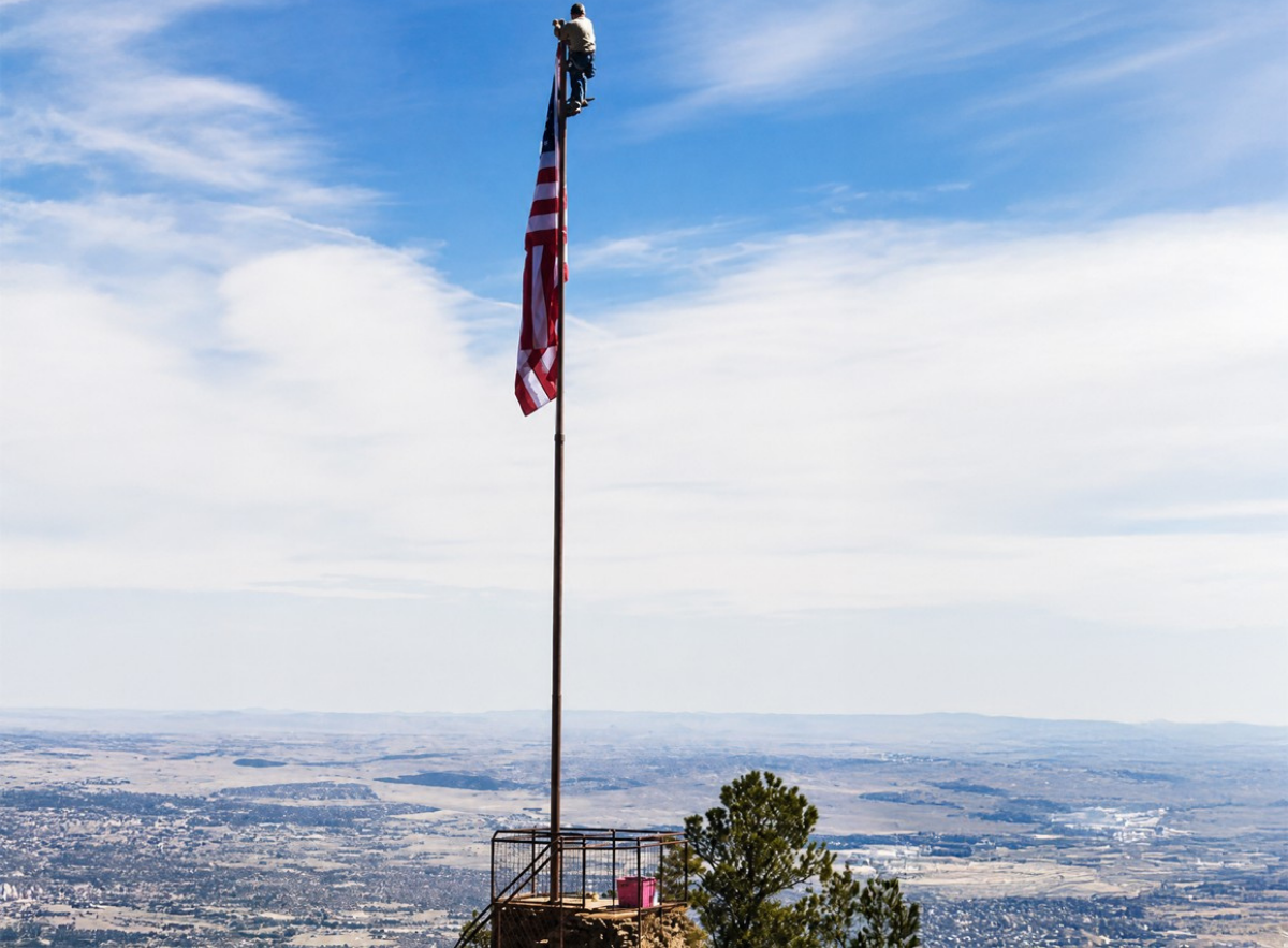 Flagpole climber at the top of a tall pole with American flag and wide landscape view